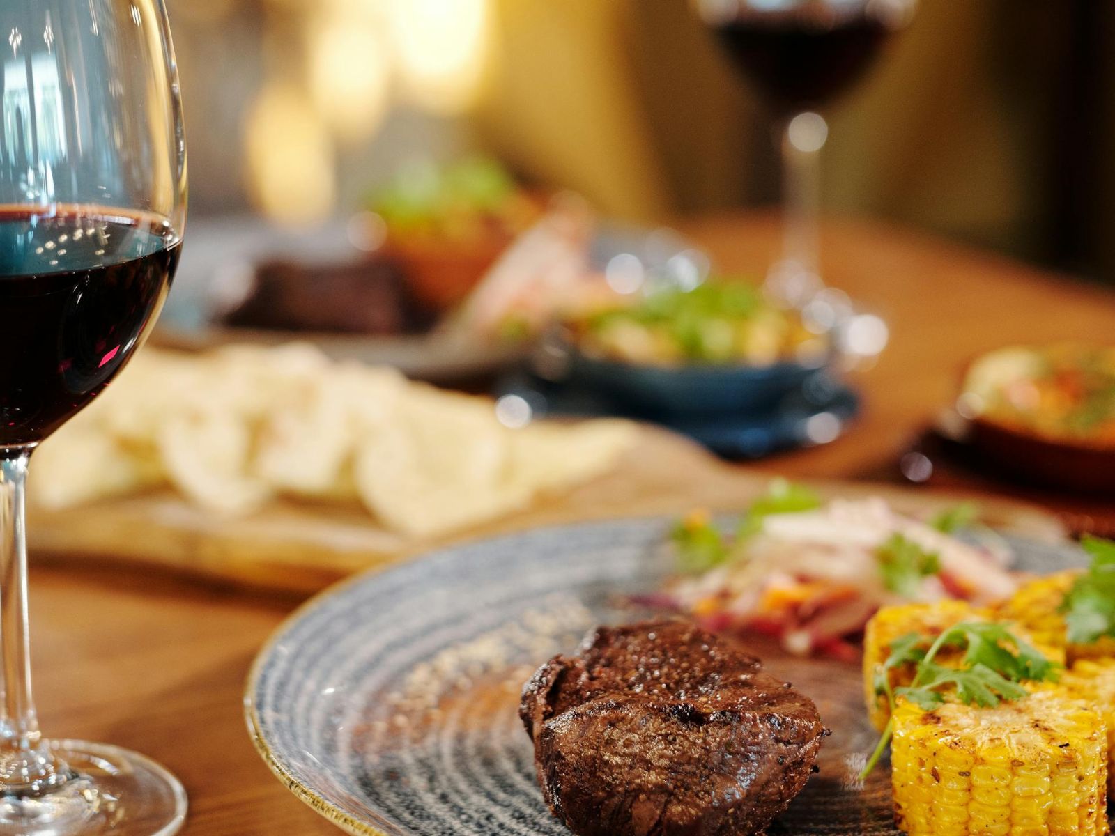 Steak dinner with corn on the cob and a glass of red wine on a candlelit table.